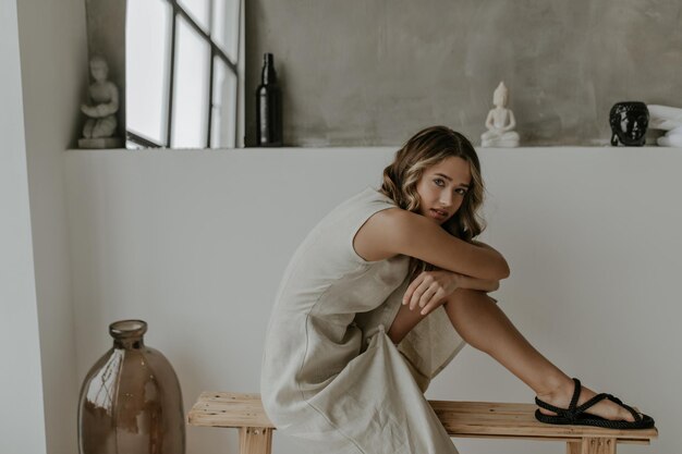 Affascinante giovane donna in elegante abito beige si siede in una posa comoda su una panca di legno e si appoggia su h