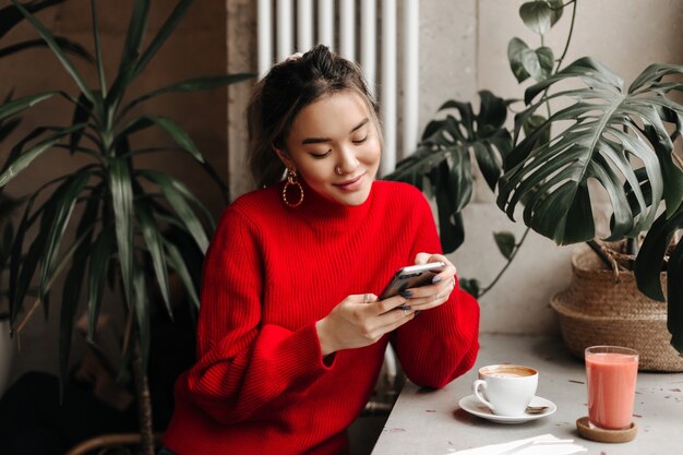 Affascinante giovane donna in enormi orecchini e maglione luminoso chat nel telefono mentre era seduto in un caffè davanti a una tazza di caffè