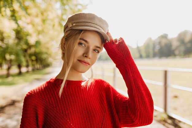 Affascinante giovane donna in pullover rosso e cappello leggero che sembra buono nel parco. Bella bionda che indossa bei accessori e vestiti alla moda in autunno.