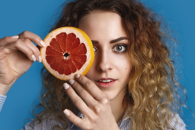 Affascinante giovane femmina carina con gli occhi verdi e capelli voluminosi in posa, nascondendo un occhio dietro il giro di succoso pompelmo maturo. Freschezza, vitamine, salute, cura, bellezza e concetto di benessere