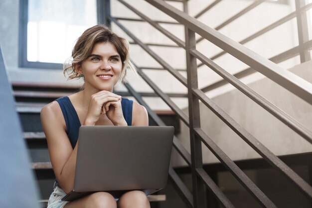 Affascinante giovane studentessa sognante sedersi all'aperto università campus scale finitura progetto sorridente sollevato sguardo felice da parte tenere laptop ginocchia inviare attività freelance lavorando nuovo programma