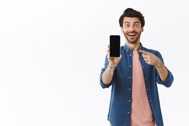 Affascinante impressionato, sorridente ragazzo felice con la barba, tenendo in mano lo smartphone, mostrando qualcosa sullo schermo vuoto, indicando il display e sorridendo stupito, consiglia una buona app, omaggio, muro bianco