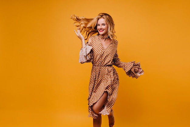 Affascinante modello caucasico in abito ballando con il sorriso. splendida ragazza bionda isolata sul muro giallo.