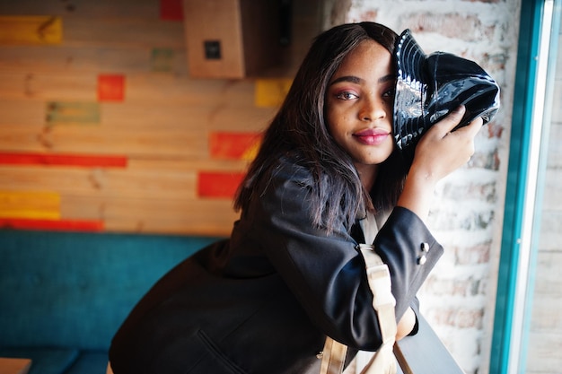 Affascinante modello di donna afroamericana in giacca nera e cappello che si rilassa nella caffetteria durante il tempo libero