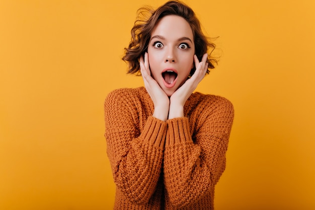 Affascinante modello femminile bianco con taglio di capelli alla moda che esprime stupore e tocca il viso