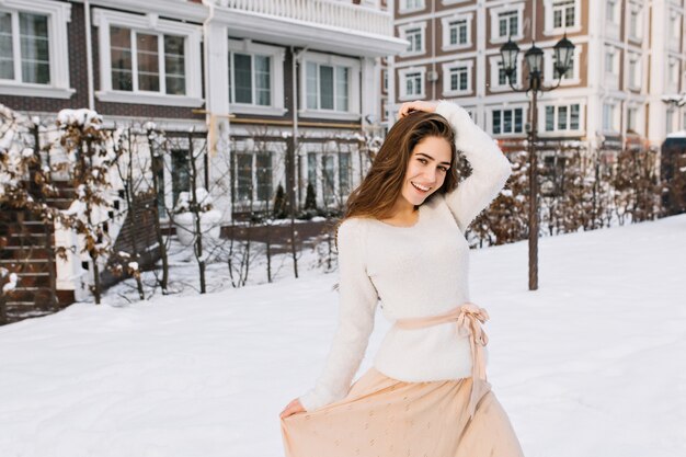 Affascinante modello femminile in maglione e gonna rosa che balla in cortile nella mattina d'inverno. Foto all'aperto di splendida donna che gode della prima neve nel giorno di dicembre.