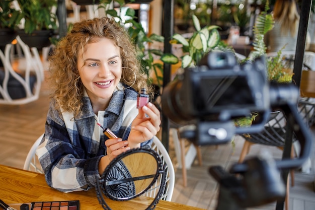 Affascinante ragazza che registra il suo episodio di video blog sui nuovi prodotti cosmetici per rossetti mentre è seduta al tavolo a casa e si trucca