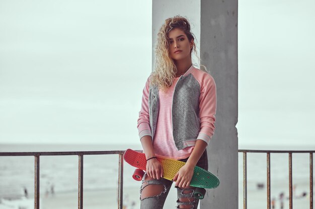 Affascinante ragazza con i capelli biondi vestita con una giacca rosa tiene uno skateboard in posa vicino a un guardrail su sfondo bianco e nero della costa del mare.
