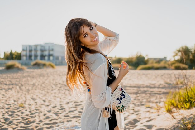 Affascinante ragazza con i capelli castani ondulati, vestita di bianco boho cover up, camminando sulla soleggiata spiaggia estiva. Concetto di viaggio e vacanza.