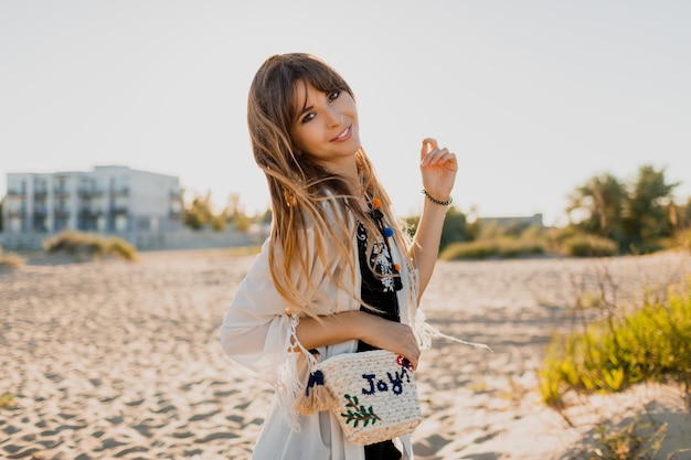 Affascinante ragazza con i capelli castani ondulati, vestita di bianco boho cover up, camminando sulla soleggiata spiaggia estiva. Concetto di viaggio e vacanza.