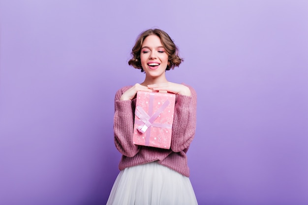 Affascinante ragazza con i capelli corti ricci in posa con scatola regalo rosa e ridendo. Modello femminile attraente con regalo di Natale isolato sul muro viola e sorridente.