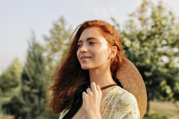 Affascinante ragazza con una lunga acconciatura allo zenzero in moderno cappello di paglia e vestito estivo alla moda che sorride distogliendo lo sguardo e posando all'aperto