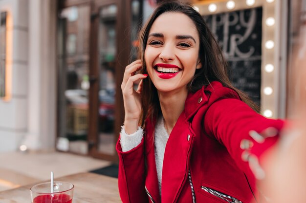 Affascinante ragazza dai capelli castani che fa selfie mentre riposa in street cafe