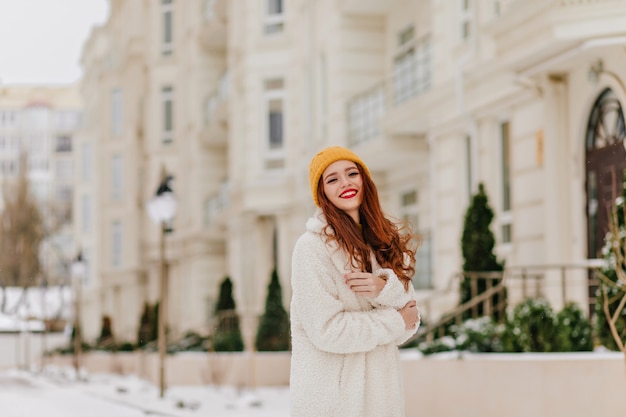 Affascinante ragazza dai capelli lunghi in posa sulla strada di sfocatura. Modello femminile attraente con i capelli dello zenzero che gode dell'inverno.