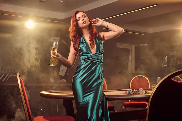 Affascinante ragazza dai capelli rossi in un lungo abito di raso blu, con un bicchiere di champagne in mano è in posa contro un tavolo da poker in un casinò di lusso. Passione, carte, fiches, alcol, vincita, gioco d'azzardo: è fem