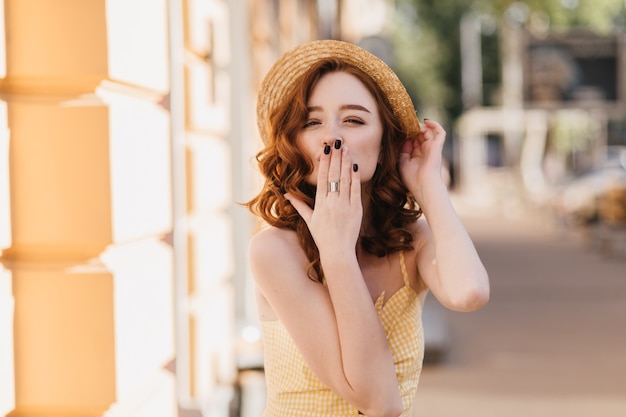 Affascinante ragazza dello zenzero in abito giallo che manda un bacio d'aria durante il servizio fotografico estivo. Colpo esterno di giocosa donna dai capelli rossi in cappello di paglia che esprime amore.