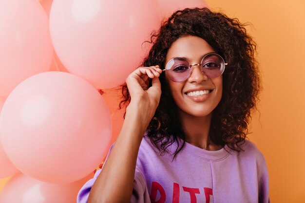 Affascinante ragazza di compleanno che tocca i suoi occhiali durante il tiro. donna africana alla moda che tiene i palloncini rosa del partito.