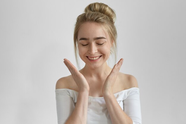 Affascinante ragazza europea con nodo di capelli che chiude gli occhi e sorride ampiamente, esprimendo gioia ed emozioni positive, tenendosi per mano davanti a lei, ricevendo presente, proposta di matrimonio o buone notizie