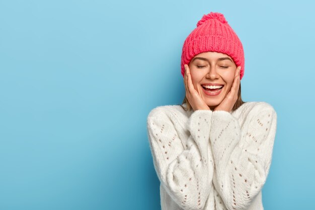 Affascinante ragazza femminile sorridente tocca le guance con entrambe le mani, gli occhi chiusi, il sorriso seducente a trentadue denti, indossa un maglione bianco invernale, posa contro il muro blu dello studio, ha una pelle pura e sana, si sente sollevato