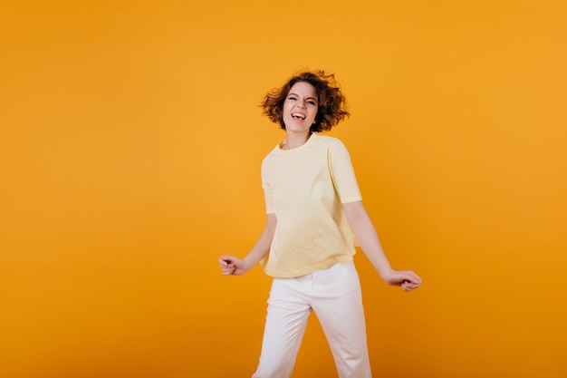 Affascinante ragazza in maglietta gialla in posa emotivamente. Ritratto dell'interno della ragazza caucasica alla moda ballando in pantaloni bianchi e facendo facce buffe.