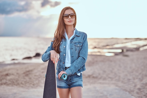 Affascinante ragazza in occhiali da sole vestita con pantaloncini di jeans e giacca in posa con uno skateboard sulla spiaggia con tempo nuvoloso durante il tramonto.