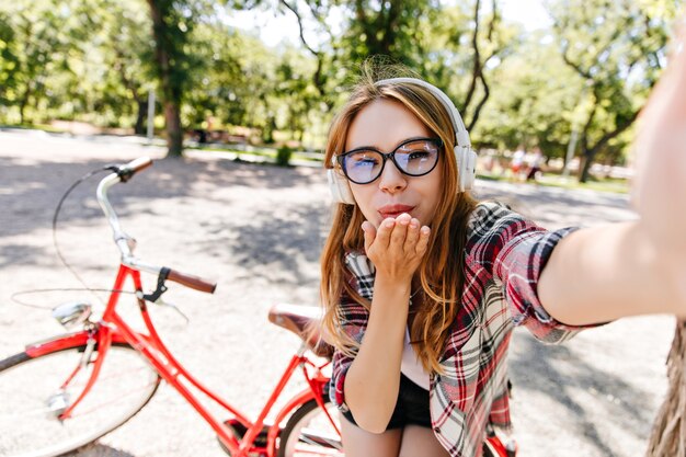 Affascinante ragazza in vetri che fanno selfie davanti alla bicicletta rossa. Colpo esterno di allegra signora europea che riposa nel parco in mattinata estiva.