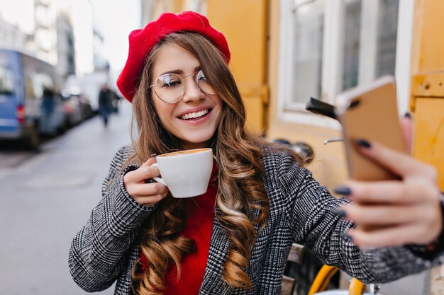 Affascinante ragazza pallida in bicchieri facendo selfie mentre beve il caffè nella caffetteria all'aperto