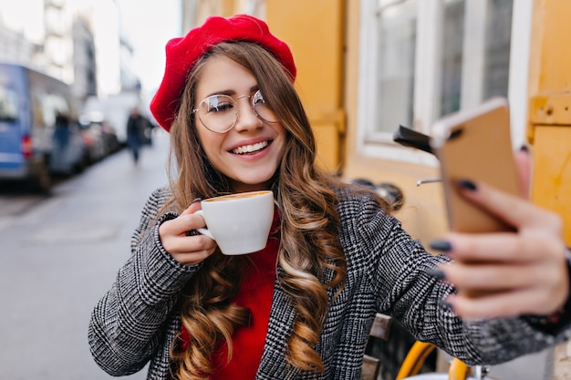 Affascinante ragazza pallida in bicchieri facendo selfie mentre beve il caffè nella caffetteria all'aperto