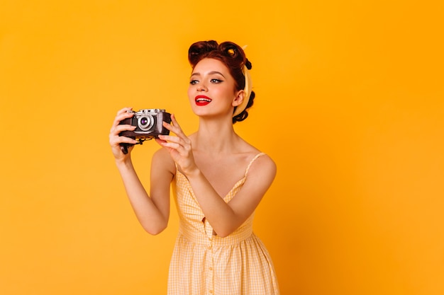 Affascinante ragazza pinup scattare foto. Ispirato la donna allo zenzero con la fotocamera in piedi sullo spazio giallo.