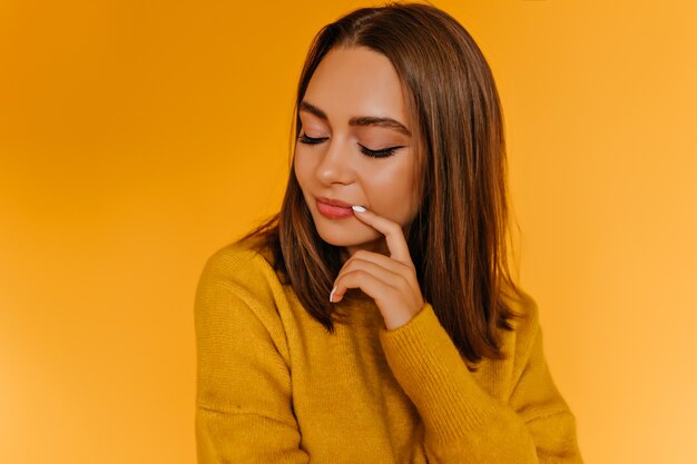 Affascinante signora con capelli lucidi in posa con gli occhi chiusi. Foto interna della ragazza con il trucco alla moda isolato sulla parete arancione.