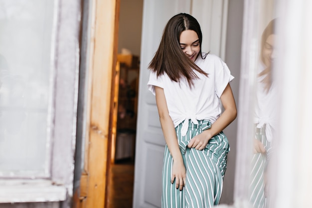 Affascinante signora europea che ride mentre scherza a casa. Ritratto dell'interno della ragazza castana spensierata in pantaloni verdi che godono del fine settimana.