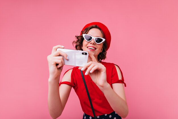 Affascinante signora francese con gli occhiali vintage che si prende una foto. donna riccia in berretto rosso che fa selfie.