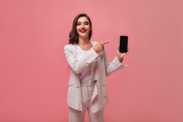 Affascinante signora in abito beige dimostra smartphone su sfondo rosa. Donna alla moda con trucco luminoso in abito bianco mostra sul suo telefono.