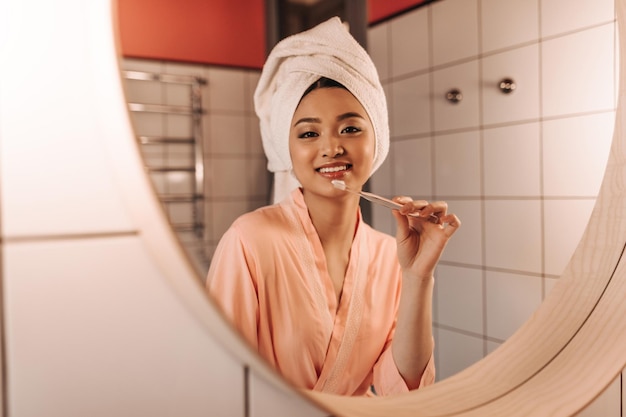 Affascinante signora in asciugamano si guarda allo specchio e si pulisce i denti in bagno