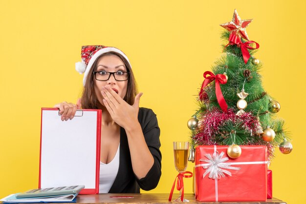 Affascinante signora in tuta con cappello di Babbo Natale e occhiali da vista che mostra il documento che si sente sorpresa in ufficio