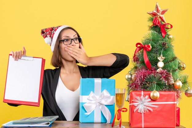 Affascinante signora in tuta con cappello di Babbo Natale e occhiali da vista in possesso di documenti sorprendentemente in ufficio