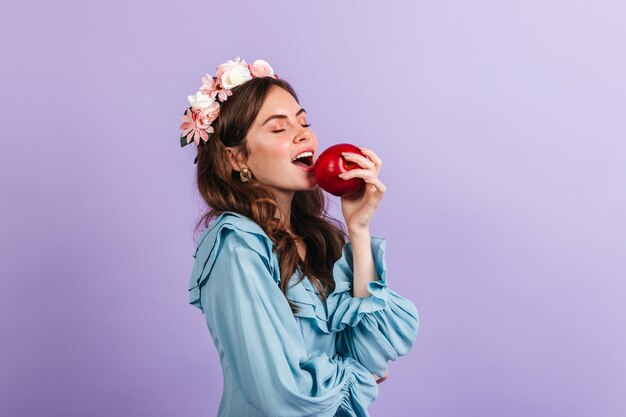 Affascinante signora in una corona di fiori morde una mela succosa. Ritratto di ragazza in camicetta blu sulla parete lilla.