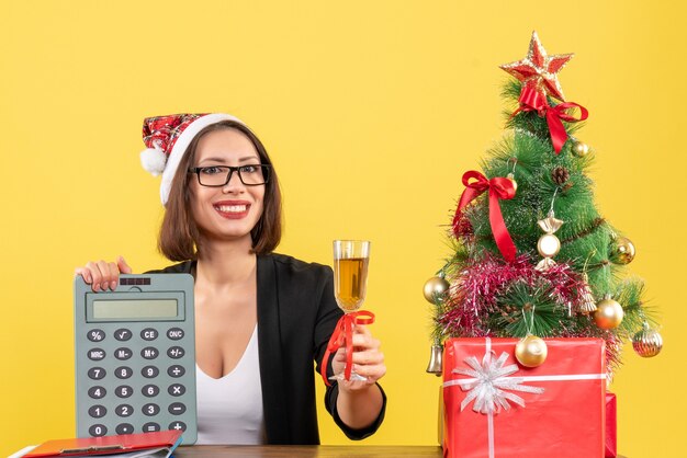 Affascinante signora sorridente in vestito con cappello di Babbo Natale e occhiali da vista mostrando calcolatrice e alzando il vino in ufficio su giallo isolato