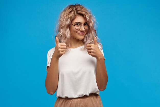 Affascinante splendida giovane donna hipster con voluminosi capelli rosati che sorride felicemente alla macchina fotografica, mostrando il pollice in alto gesto