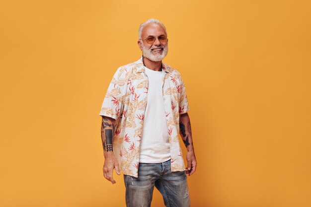 Affascinante uomo in jeans, camicia e occhiali da sole posa sul muro arancione