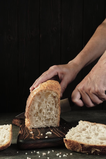 Affettare il pane su una tavola di legno isolata su uno sfondo scuro La donna taglia il pane fresco artigianale sul telaio verticale del tavolo della cucina Cibo sano e concetto di panetteria tradizionale