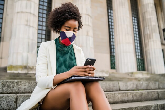 Afro business donna che indossa la maschera protettiva e utilizzando il suo telefono cellulare mentre era seduto sulle scale all'aperto in strada. Business e concetto urbano.