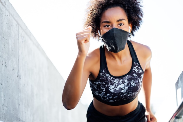 Afro donna atletica che indossa la maschera per il viso durante l'esecuzione all'aperto. Nuovo stile di vita normale. Sport e concetto di stile di vita sano.