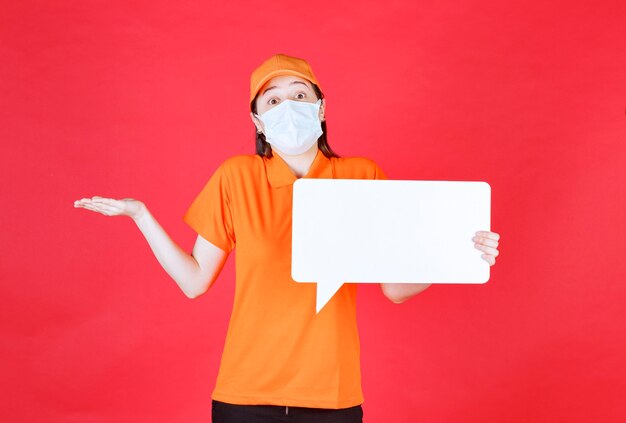 Agente di servizio femminile in codice di abbigliamento di colore arancione e maschera con in mano una bacheca informativa rettangolare bianca e sembra confusa e incerta