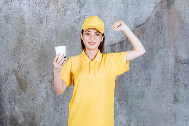 Agente di servizio femminile in uniforme gialla che tiene un bicchiere di plastica e che mostra il segno positivo della mano.