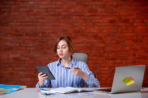 Agente di viaggio vista frontale seduto dietro il suo posto di lavoro con laptop e blocco note all'interno mappa agenzia servizio assistente biglietto mondo globale