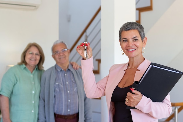 Agente immobiliare femminile che dà le chiavi alle coppie anziane. Famiglia sorridente con gli occhiali in piedi accanto alla commessa in abito rosa. Immobiliare, concetto di acquisto