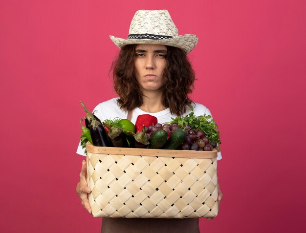 Aggrottando le sopracciglia giovane giardiniere femminile in uniforme che indossa il cappello da giardinaggio tenendo il cesto di verdure