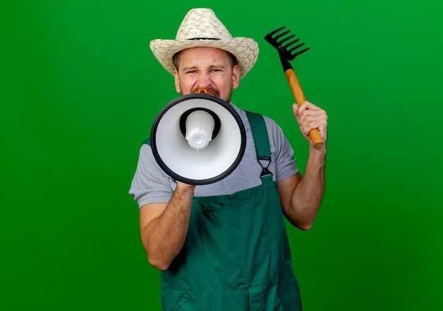 Aggrottando le sopracciglia giovane giardiniere slavo bello in uniforme e cappello guardando tenendo il rastrello e parlando da altoparlante