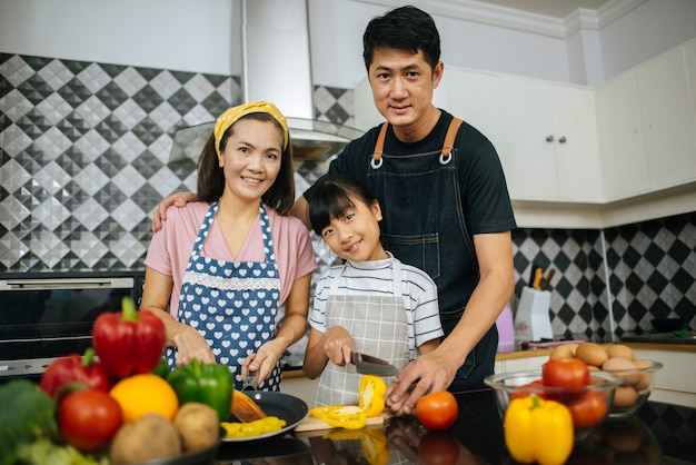 Aiuto sveglio della ragazza i suoi genitori stanno tagliando le verdure e stanno sorridendo mentre cucinano insieme nella cucina